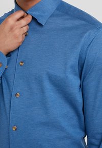 Camicia blu con bottoni realizzata in tessuto texturizzato. Presenta un colletto appuntito e bottoni marroni. Primo piano del colletto e del torso superiore visibili.