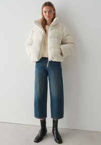 Giacca in fleece color crema, morbida e pelosa, con colletto alto e chiusura con zip, abbinata a jeans a gamba larga blu scuro e stivaletti neri alla caviglia.