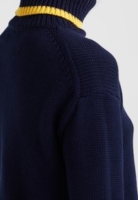 Pull en tricot marine avec un col montant doté d'un bord côtelé jaune contrastant et des détails texturés sur les coutures des épaules.