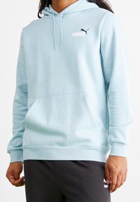 Vaaleansininen huppari pehmeää kangasta, jossa edessä tasku, resorit hihansuissa ja pieni musta Puma-logo rinnassa.