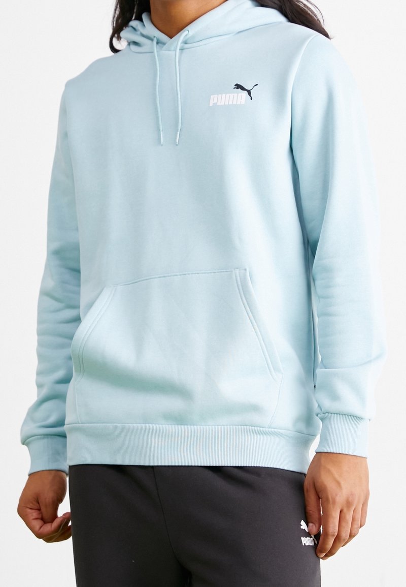 Vaaleansininen huppari pehmeää kangasta, jossa edessä tasku, resorit hihansuissa ja pieni musta Puma-logo rinnassa.