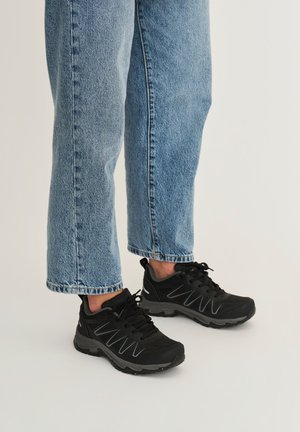 Persoon die een blauwe spijkerbroek en zwarte sportieve sneakers met dikke zolen draagt, staand op een lichte achtergrond.