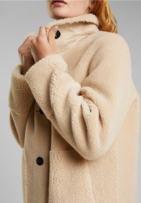 Beige fleece-kappa med hög krage, svarta knappar och textil med struktur, med vida ärmar och avslappnad passform.