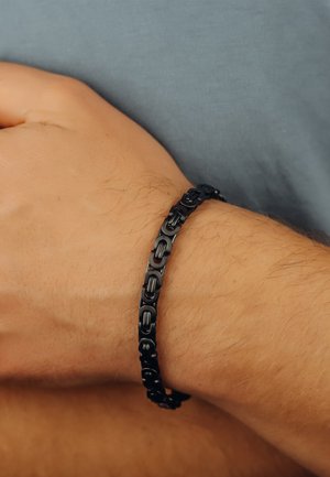 Schwarzes Armband aus ineinander verwobenen Metallgliedern mit glatter Oberfläche, am Handgelenk getragen vor einem grauen Hintergrund.