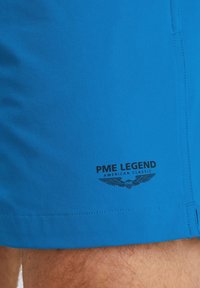 Plave kupaćice od lagane tkanine s glatkom teksturom. Imaju crni logo na donjem lijevom dijelu s dizajnom grba.