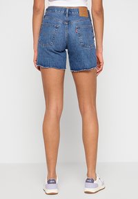 Vue arrière d’une personne portant un short en jean bleu à ourlets effilochés et des baskets blanches Levi’s sur fond uni.