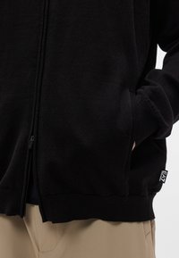 Μαύρο φούτερ με φερμουάρ, κατασκευασμένο από μαλακό ύφασμα, με ψηλό γιακά, ριμπ μανσέτες, πλαϊνές τσέπες και ετικέτα κοντά στο τελείωμα.