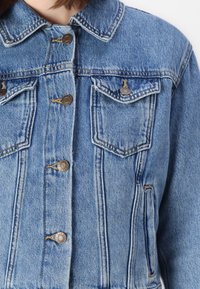 Ανοιχτού μπλε τζιν μπουφάν με κουμπωτή μπροστινή πλευρά, δύο τσέπες στο στήθος και κλασικό γιακά. Υφασμάτινη επιφάνεια με ορατές λεπτομέρειες ραφής.