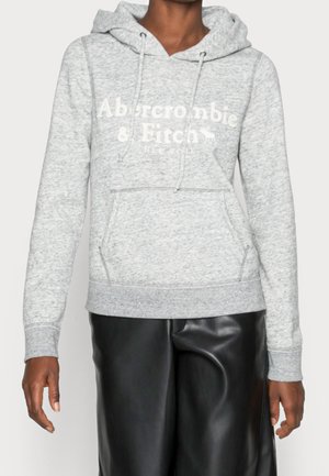 Persona che indossa un hoodie grigio chiaro di Abercrombie & Fitch con tasca frontale e pantaloni in pelle nera, in piedi contro uno sfondo semplice.