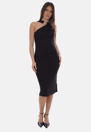 Donna che indossa un abito midi nero aderente senza maniche con una spallina singola e dettaglio ad anello dorato, abbinato a décolleté nere con punta appuntita.