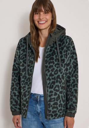 Kvinde smilende iført en grøn og sort leopardprint hættetrøje med lynlås og snører, en hvid skjorte og blå jeans mod en lys baggrund.