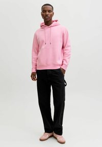 Sweat à capuche rose avec poche avant et cordon de serrage, associé à un pantalon noir et des baskets rose clair avec des accents blancs. Texture de tissu lisse.