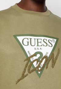 Sudadera verde oliva con un logo triangular blanco que incluye el texto "GUESS" y "U.S.A.", acentuada con un bordado texturizado en oro que reza "ICON".