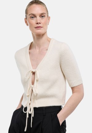 Femme portant un cardigan en maille crème à manches courtes avec deux liens à l'avant, par-dessus un pantalon noir, debout les mains dans les poches devant un fond blanc.
