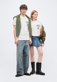 Νεαρός άνδρας και γυναίκα φορούν λευκά T-shirt με σχέδια· ο άνδρας έχει πράσινο πουλόβερ περασμένο στους ώμους και φαρδιά τζιν, η γυναίκα φοράει τζιν φούστα και μαύρες μπότες.
