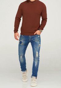 Brun stickad tröja, slitna blå jeans med hål, och beige sneakers med remmar. Avslappnad outfit med lös passform.