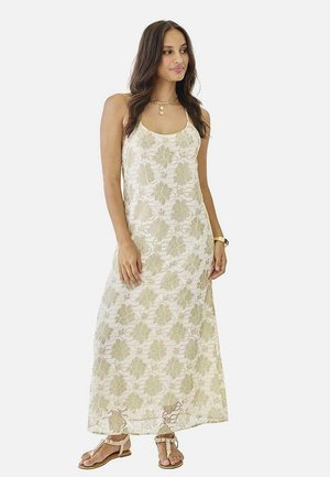 Mujer vestida con un vestido maxi blanco sin mangas con estampados florales beige, combinada con sandalias, un collar y un reloj de pulsera, de pie y mirando hacia la derecha.