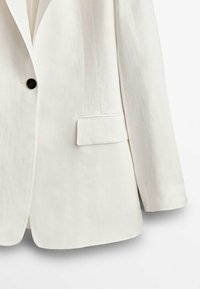 Valkoinen yksitakkinen bleiseri, jossa on aansäkkilapeli, pitkät hihat ja läpällinen tasku oikealla puolella.