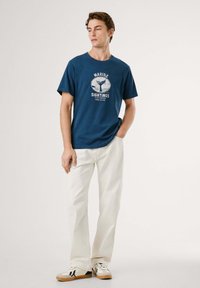 Camiseta de algodón azul marino con un estampado gráfico de temática marina. Combinada con pantalones rectos blancos y zapatillas casuales con detalles en negro.