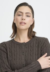 Młoda kobieta o prostych brązowych włosach, ubrana w ciemnobrązowy sweter na guziki z warkoczowym wzorem, ze skrzyżowanymi rękami, na jasnym neutralnym tle.