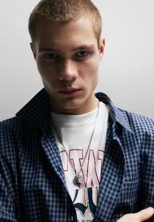 Kockás kék-fehér gombos ing, fehér, grafikás póló fölött rétegezve, ezüst lánc medállal. Semleges hátterű.