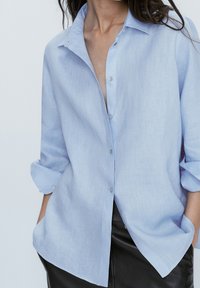 Camicia in lino azzurro chiaro con vestibilità rilassata, colletto appuntito e chiusura a bottoni; presenta maniche arrotolate e un tessuto morbido e testurizzato.