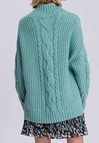 Pull tricoté turquoise avec un col montant, des poignets côtelés, des motifs texturés et un dessin en torsade au centre. Conception en maille douce et épaisse.