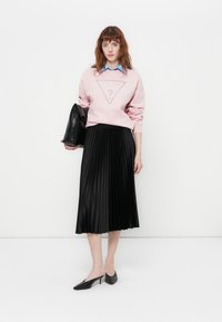 Mujer vestida con sudadera rosa sobre camisa de cuello azul, falda midi plisada negra, tacones negros, sosteniendo un gran clutch negro frente a un fondo blanco.