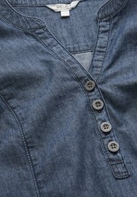Dunkelblaue Jeanshemd mit V-Ausschnitt, ausgestattet mit fünf schwarzen Knöpfen, subtilen Nähten und einer aufgesetzten Tasche auf der linken Seite.