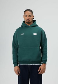 Groene oversized hoodie gemaakt van zachte stof, met een voorzak, logopatches en geribbelde manchetten. Draagt donkere jeans.