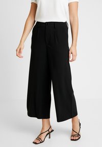 Zwarte wijde pantalon met een gladde textuur, voorplooien en een knoopsluiting. Gecombineerd met zwarte sandalen met banden.