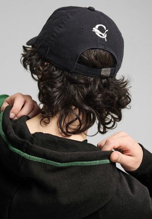 Czarna czapka baseballowa z logo z tyłu, noszona przez osobę z kręconymi włosami. Ciemna bluza z kapturem z widocznymi zielonymi akcentami w szyciu.