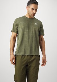Oliven grøn kortærmet t-shirt med et struktureret camouflagemønster og sideslids. Har et hvidt Under Armour-logo på brystet.