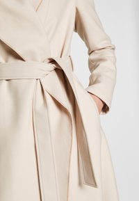 Abrigo de lana beige con un amplio cuello y cinturón a juego. La tela es suave con una textura sutil y un diseño limpio y estilizado.