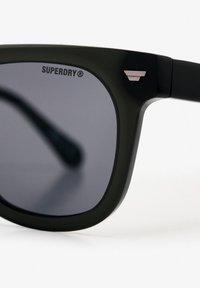 Matte zwarte zonnebril met donkere glazen, voorzien van een klein metalen accent en het "SUPERDRY" logo gedrukt op het bovenste montuur.