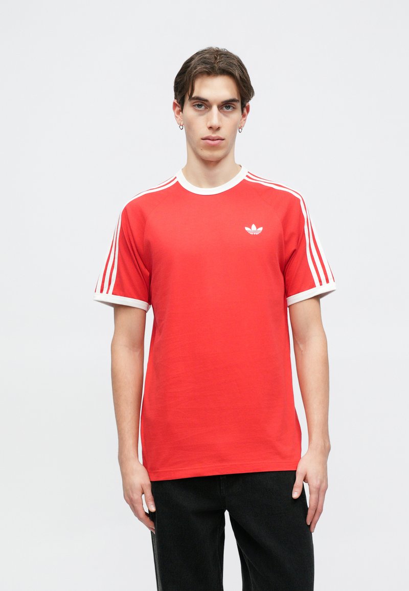 T-shirt in cotone rosso con colletto e polsini a coste bianchi. Presenta tre strisce bianche sulle spalle e un piccolo logo bianco sul petto.