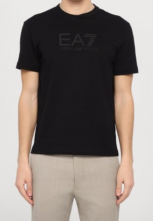 Uomo che indossa una maglietta nera con il logo Emporio Armani EA7 e pantaloni beige, visibile dal collo ai fianchi su sfondo semplice.