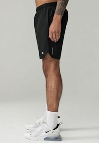 Black athletic shorts with an elastic waistband, side slits for mobility, and a reflective logo detail on the hem. Worn with white sneakers.
