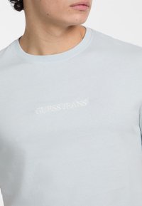 Gaiši zils kokvilnas T-krekls ar apaļu kakla izgriezumu, ar izšūtu "GUESS JEANS" logo baltā krāsā uz krūšu zonas.
