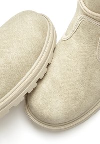 Beige Knöchelschuhe aus strukturiertem Stoff mit einer abgerundeten Zehe, dicker Gummisohle und verstärkten Nähten entlang der Kanten.