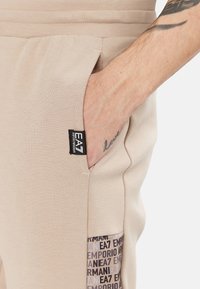 Joggers beige realizzati in tessuto morbido, dotati di tasche laterali e una tasca decorata con il marchio "EA7 Emporio Armani" sulla gamba inferiore.