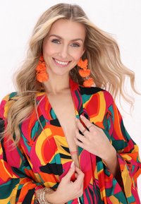 Kolorowy kimono z odważnymi wzorami w czerwieni, zieleni i pomarańczy; luźny krój z szerokimi rękawami. Pomarańczowe kolczyki z frędzlami i warstwowe bransoletki.