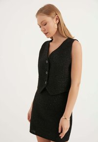 Svart tweedväst med V-hals, ärmlös design och tre knappar. Slät yta med subtila glittrande detaljer. Matchas med en passande kjol.