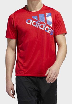 Mann trägt rotes Adidas-T-Shirt mit großem blau-weißem Logo auf der Brust und schwarze Shorts, steht vor einem einfarbigen Hintergrund.
