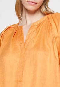 Gros plan sur une femme portant un chemisier orange ample avec un col fendu et des détails de tissu froncé sur les épaules.