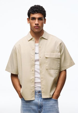 Młody mężczyzna z kręconymi włosami nosi beżową koszulę z krótkimi rękawami zapinaną na guziki, pod którą ma biały siateczkowy top oraz niebieskie dżinsy, pozując z rękami w kieszeniach.