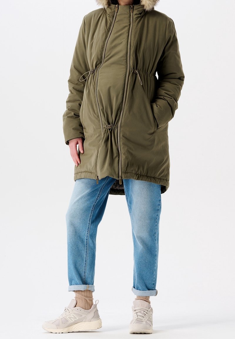 Personne portant un manteau d'hiver olive avec capuche en fourrure, un jean bleu clair retroussé, des chaussettes beiges et des baskets blanches se tenant sur fond blanc.