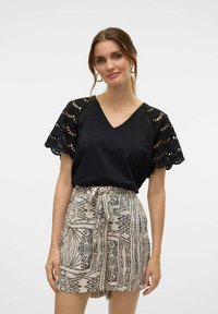Top negro de pico con intrincadas mangas cortas de encaje, combinado con pantalones cortos beige con patrones que presentan diseños geométricos negros y una cintura con lazo.