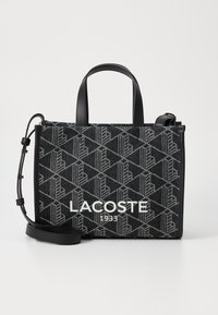 Torba na ramię w czarno-szare wzory z skórzanymi uchwytami, z wyraźnym napisem "LACOSTE 1933" z przodu. W zestawie regulowany pasek na ramię.