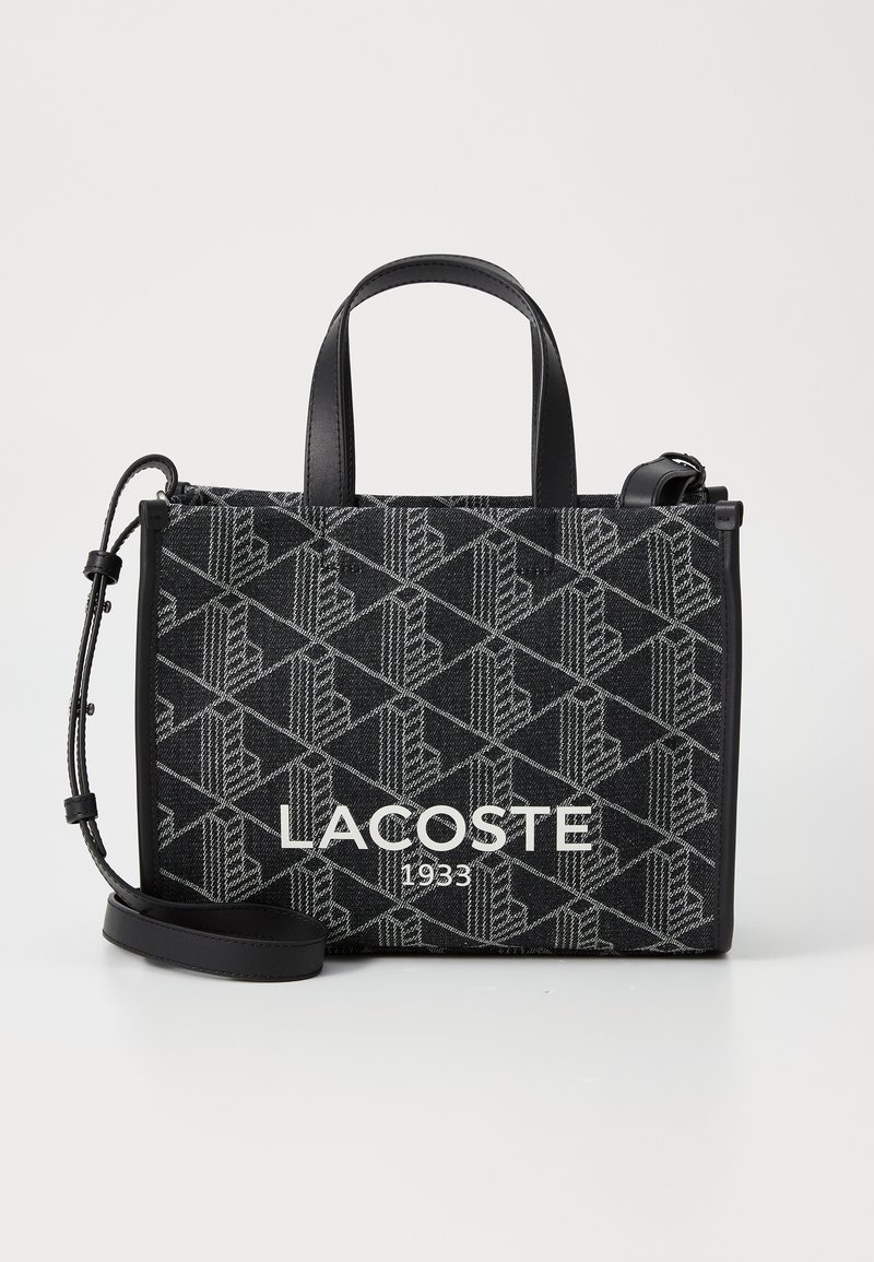 Torba na ramię w czarno-szare wzory z skórzanymi uchwytami, z wyraźnym napisem "LACOSTE 1933" z przodu. W zestawie regulowany pasek na ramię.